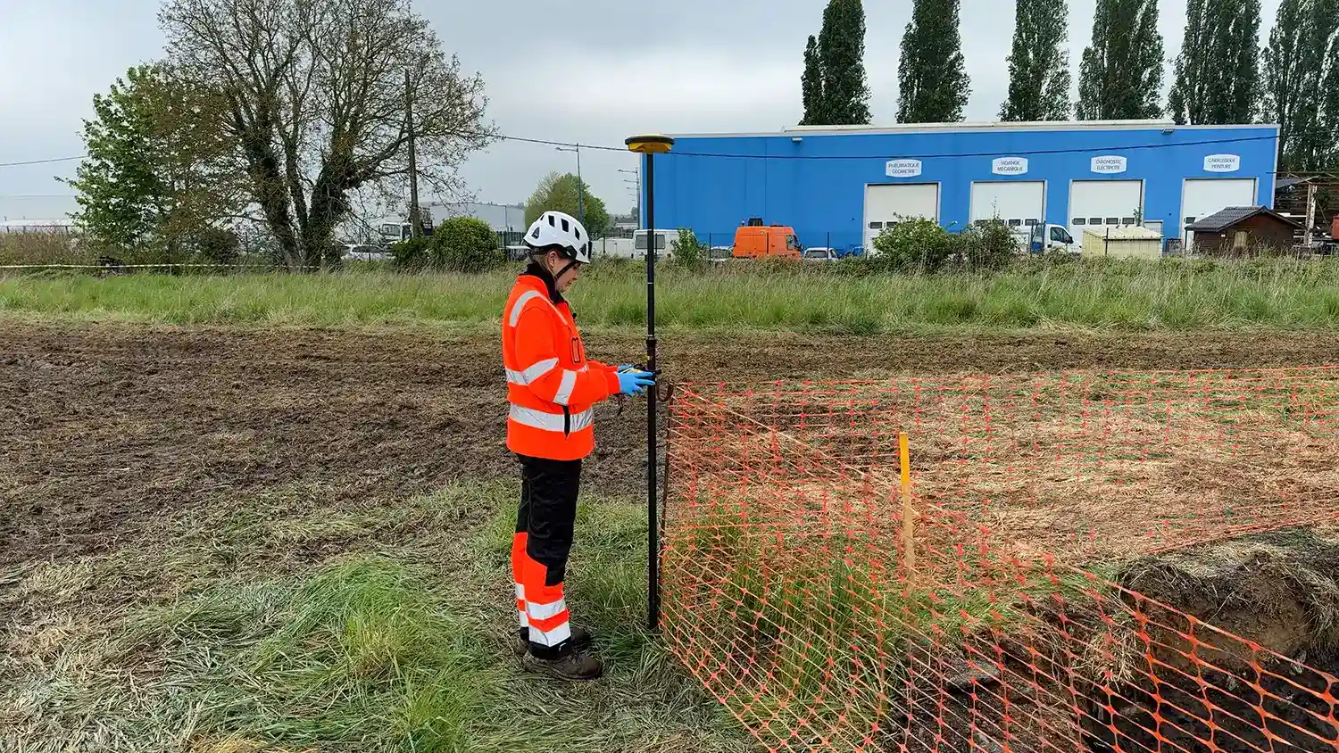 Personne en tenue orange et casque blanc effectuant des mesures près d'une clôture orange sur un site herbeux, bâtiment bleu en arrière-plan.