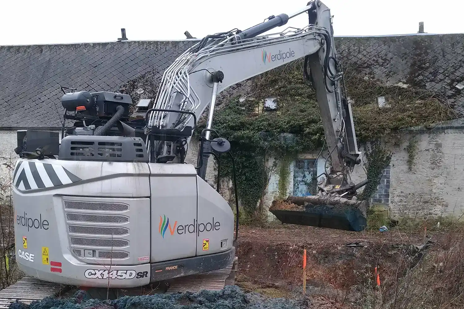 Une pelleteuse Verdiopole grise opère devant un bâtiment ancien couvert de lierre, avec des piquets orange délimitant la zone.
