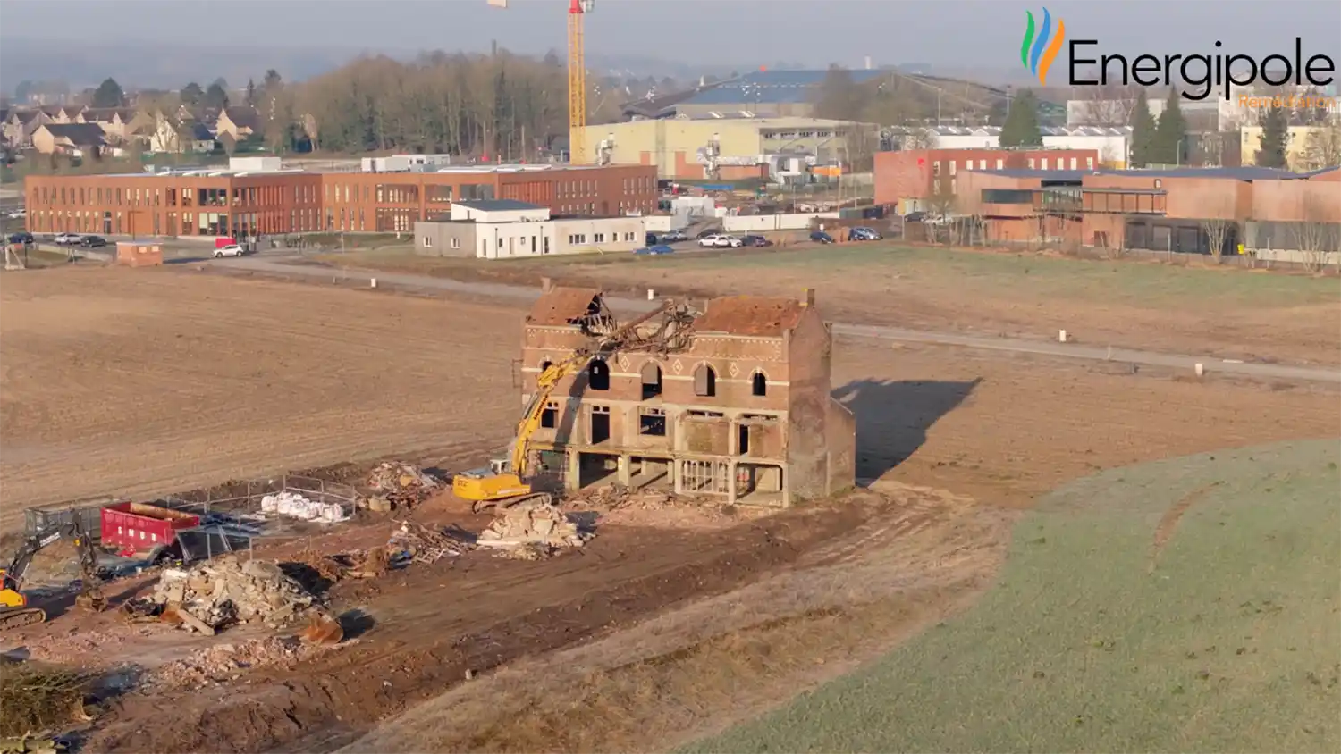 Une pelleteuse jaune démolit une brasserie en briques à Liévin, entourée de champs et de bâtiments industriels en arrière-plan.