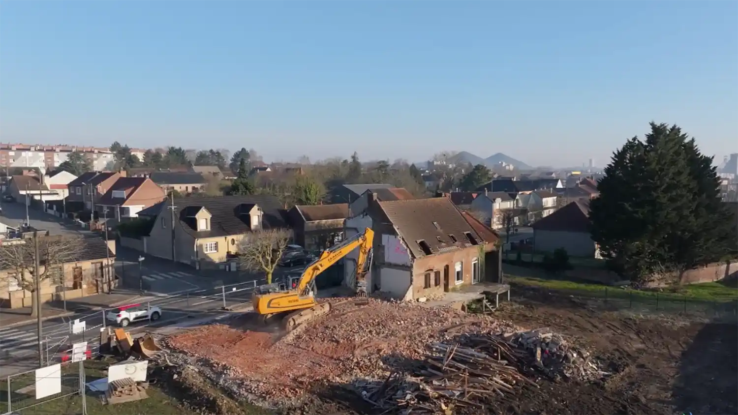 Une pelleteuse jaune démolit une maison en briques dans un quartier résidentiel à Liévin, avec des maisons et arbres en arrière-plan.