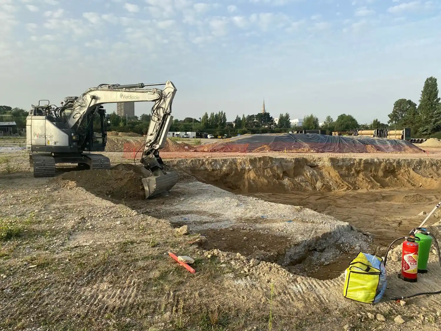 Pelleteuse grise sur un chantier de construction à Rezé, entourée de terre et de matériel de sécurité, avec un paysage urbain en arrière-plan.