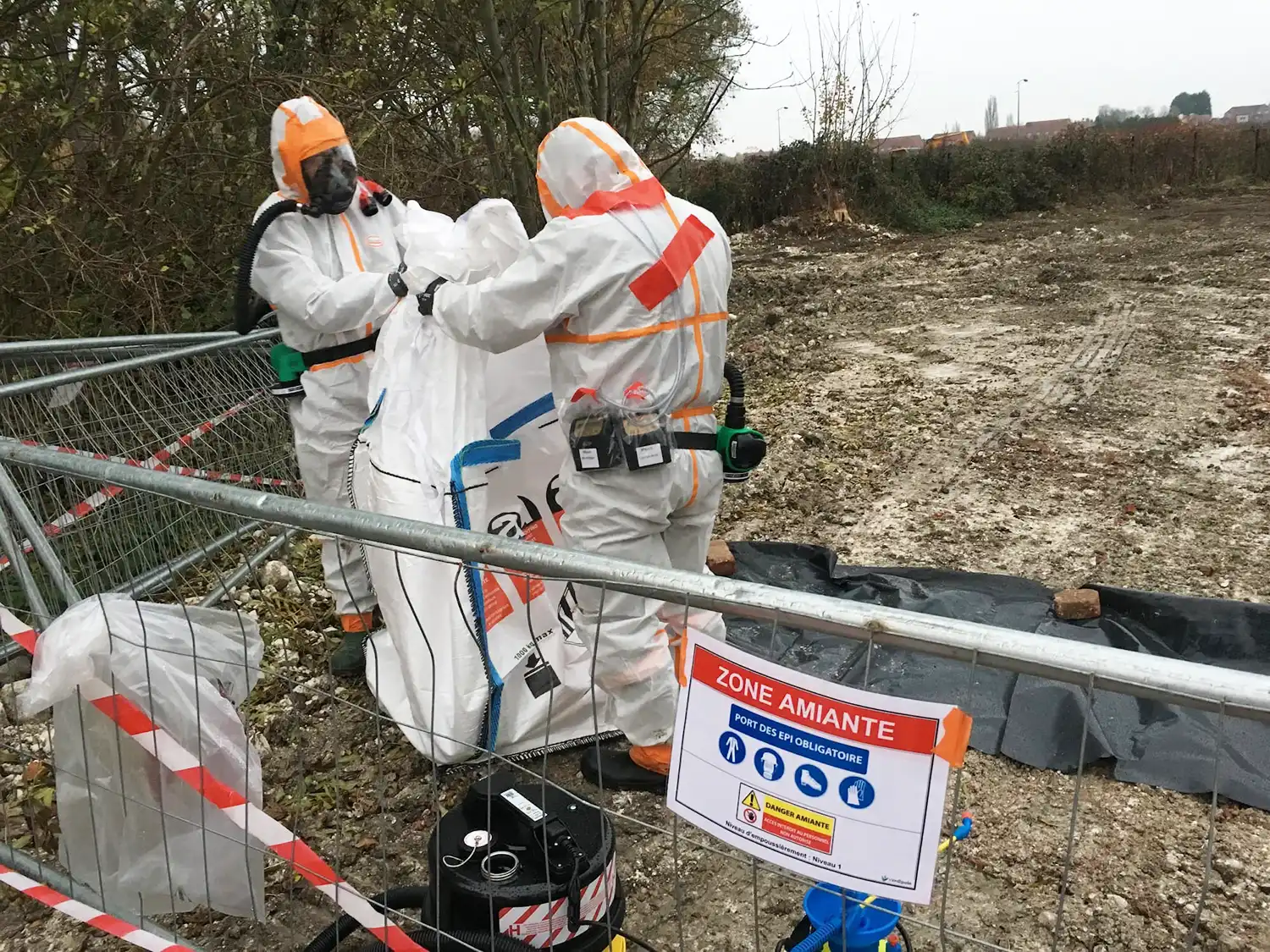 Deux personnes en combinaisons blanches et orange manipulent un sac dans une zone marquée "amiante", entourée de barrières de sécurité.