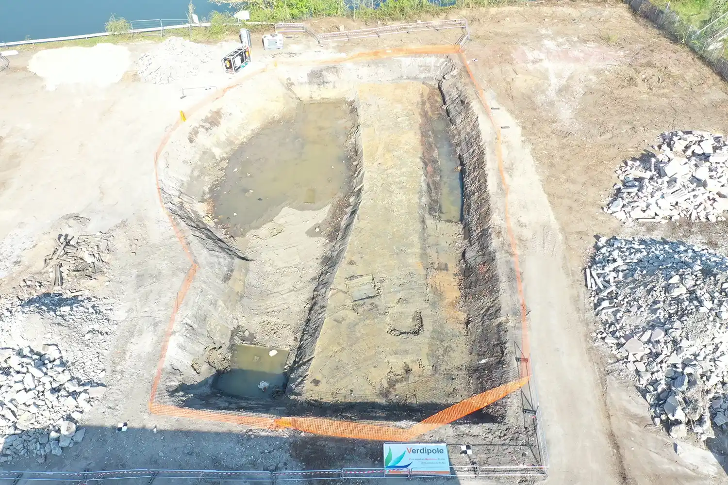 Une excavation rectangulaire remplie partiellement d'eau, entourée de barrières et de matériaux de construction.