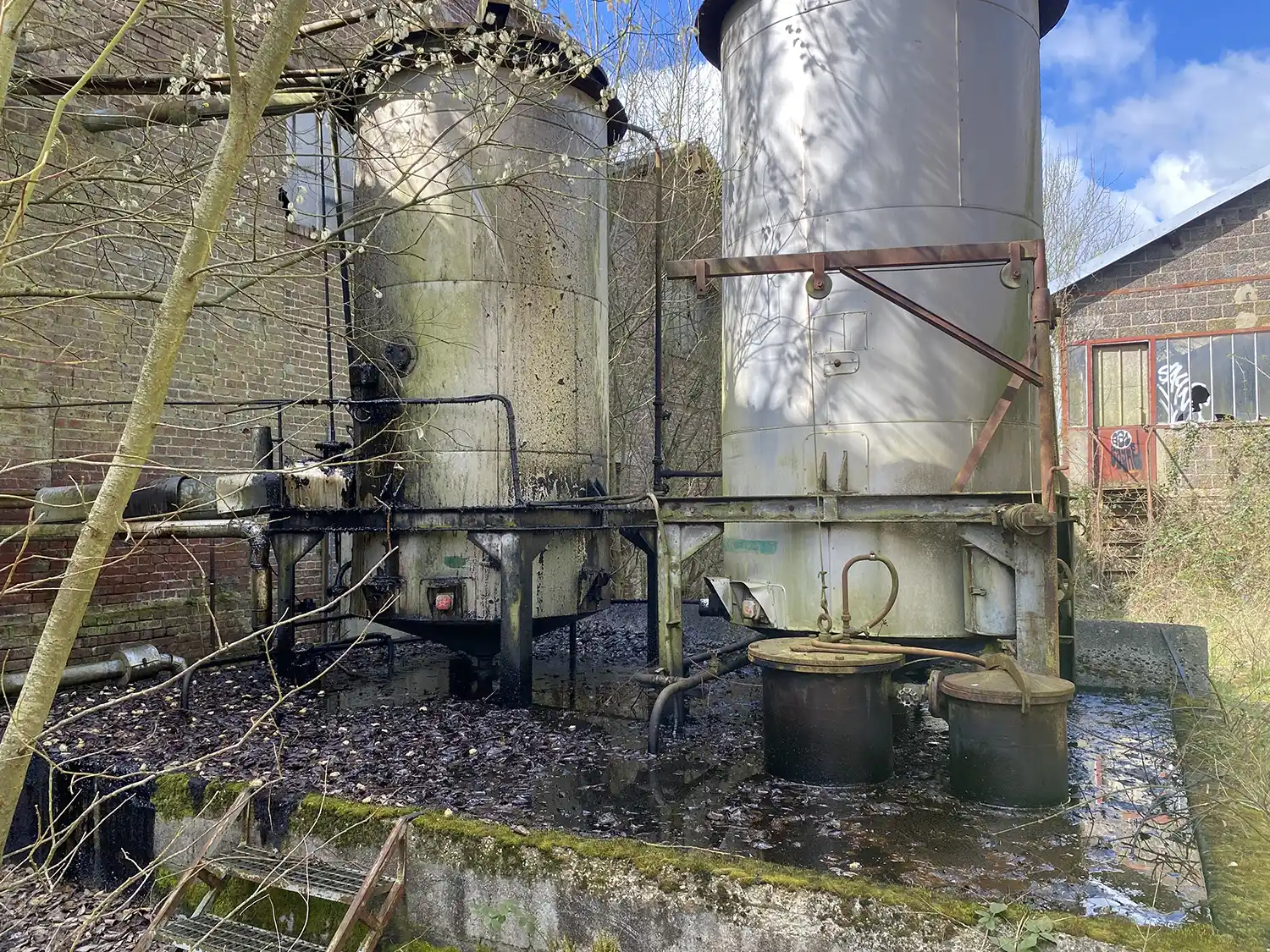 Deux grands réservoirs métalliques industriels entourés de végétation se tiennent devant un bâtiment en brique avec une fenêtre brisée.