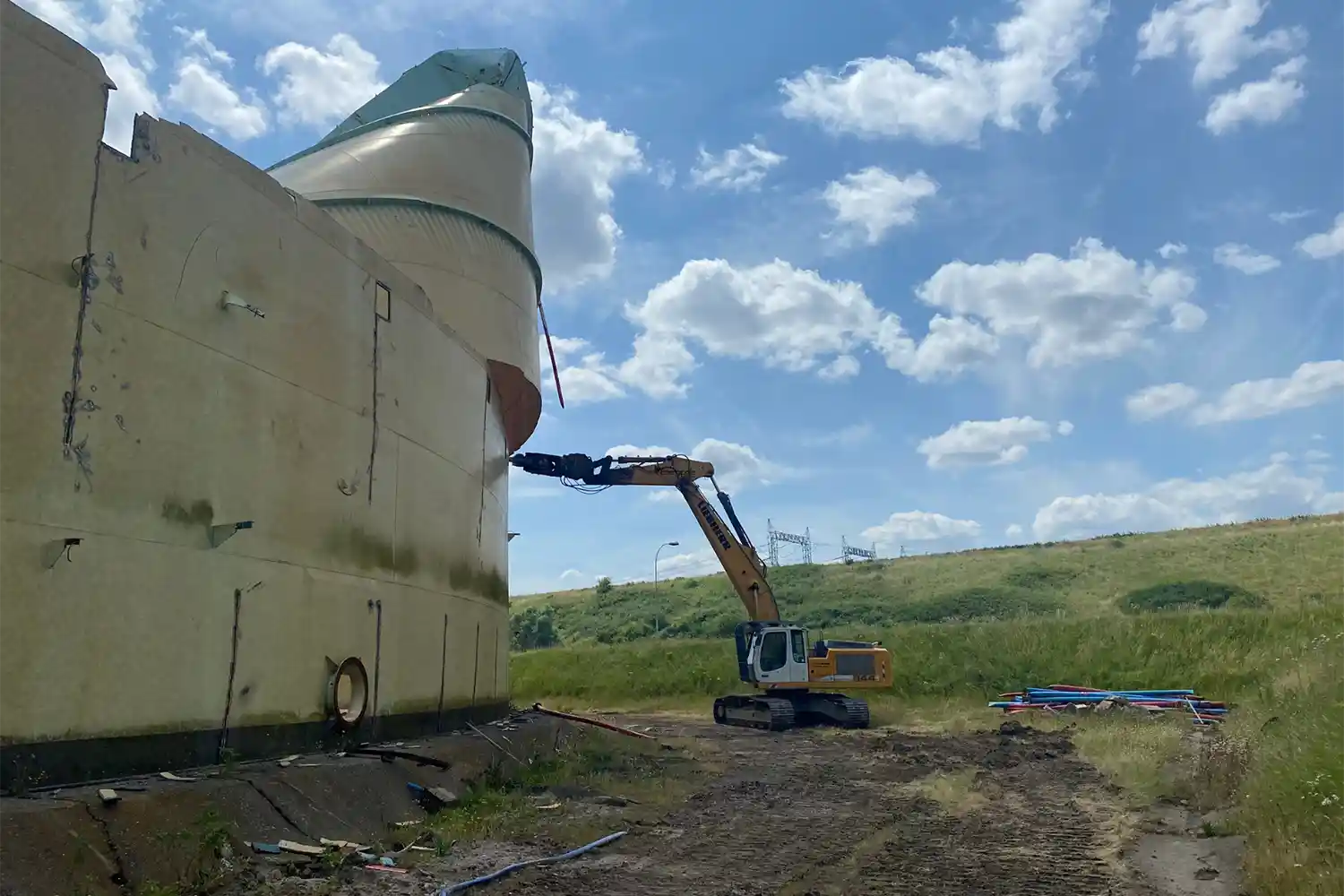 Une pelleteuse démolit une grande structure beige dans un paysage herbeux sous un ciel bleu partiellement nuageux.