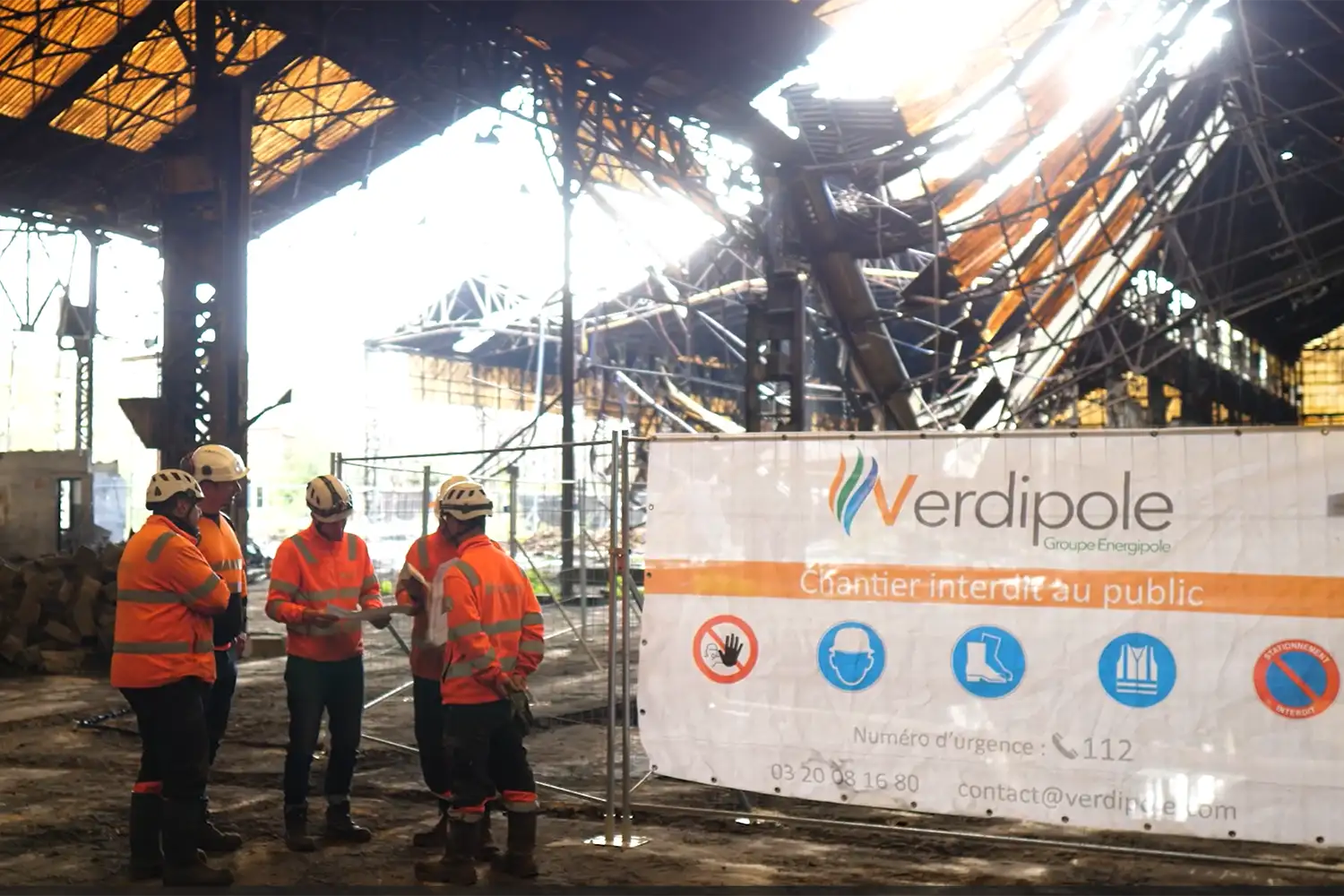 Des travailleurs en gilets orange se tiennent devant une structure métallique avec une banderole "Verdipole, chantier interdit au public".